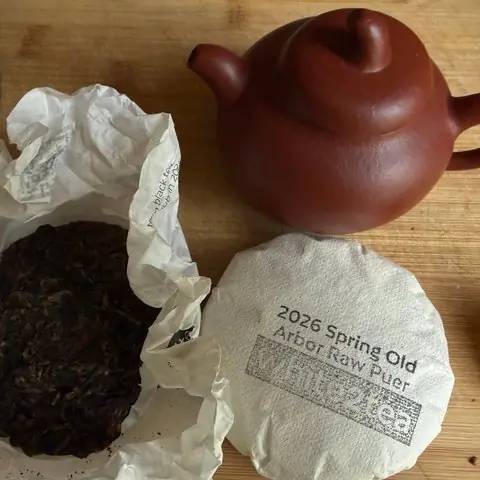 Wrapped white2tea 2026 Spring Old Arbor Raw Puer cake beside a red clay teapot and an opened tea cake on a wooden board